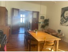 Wohnzimmer mit Essbereich in einem Altbau, erkennbar an einem Holztisch mit Stühlen, Dielenboden und hohen Decken. Im Hintergrund sind geöffnete Flügeltüren, die zu einem weiteren Raum mit großen Fenstern führen; links steht eine Küchenzeile, rechts ein Sofa und Pflanzen.