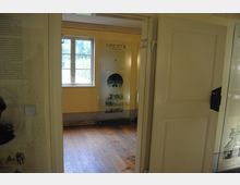 Ein Blick durch eine geöffnete Holztür in ein schlichtes Innenraum mit gelben Wänden und einem Holzboden. An der Wand hängt eine Infotafel mit Text und Bildern, ein Fenster mit Blick nach draußen lässt Tageslicht herein.