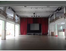 Großer, leerer Veranstaltungsraum mit einer Bühne, rotem Vorhang und einer Projektionsfläche. Der Saal hat eine hohe Decke, umlaufende Emporen mit Geländern und helle Wände.