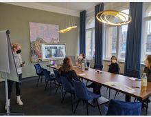 Ein moderner Konferenzraum mit einem langen Holztisch und blauen Stühlen, an dem mehrere Personen mit Masken sitzen. Im Hintergrund große Fenster mit blauen Vorhängen, eine geflieste Wand mit Kunstwerk und ein Flipchart sowie ein Bildschirm mit Videokonferenz-Schaltung.