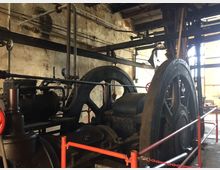 Ein alter Maschinenraum mit großen Schwungrädern und Dampfkesselanlagen, umgeben von rostigen Rohren und einer verwitterten, grob verputzten Wand. Der Raum ist spärlich beleuchtet, und die Maschinen sind durch rote Sicherheitsabsperrungen gesichert.