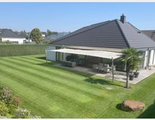 Modernes Einfamilienhaus mit flachem Satteldach, großer Terrasse mit Sonnensegel und gepflegtem Rasen im Vordergrund. Im Garten stehen ein Palmenbaum, eine Hecke als Sichtschutz, sowie ein Blumenbeet und ein dekorativer Stein.