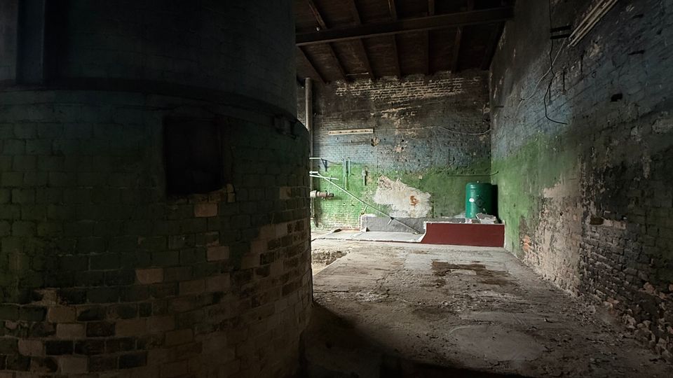 Dunkler, leerer Industrieraum mit großen zylindrischen Mauern, freiliegenden Holzbalken an der Decke und abgenutzten Ziegelwänden mit grünem Anstrich. Im Hintergrund sind Rohrleitungen, eine kleine erhöhte Plattform und ein grüner Behälter auf einem staubigen Betonboden zu sehen.