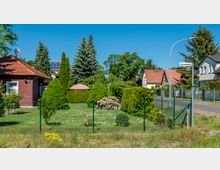 Ein gepflegter Vorgarten mit Hecken, einem Blütenstrauch und einem Sonnenschirm, umgeben von einem Metallzaun. Im Hintergrund sind Einfamilienhäuser mit roten und grauen Dächern sowie zahlreiche Bäume zu sehen; ein Straßenschild mit der Aufschrift "Kastanienallee" steht an einer Gehwegkreuzung.