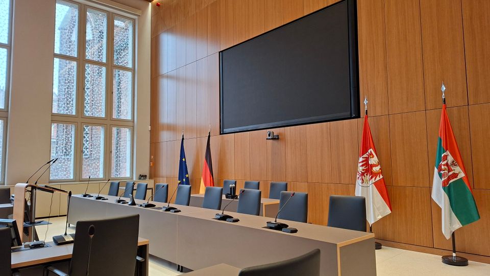 Moderner Konferenz- oder Sitzungssaal mit langen Tischen, grauen Stühlen und integrierten Mikrofonen, vor einer holzvertäfelten Wand mit großem Bildschirm. Seitlich stehen hohe Fenster, und neben dem Podium sind mehrere Flaggen (EU, Deutschland sowie Landesflaggen) aufgestellt.