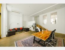 Modernes, helles Wohnzimmer mit offener Gestaltung, bestehend aus einer Sitzecke mit zwei braunen Ledersofas, einem Holztisch mit schwarzen Bürostühlen auf einem Teppich und einer kleinen Küchenzeile. Im Hintergrund führen Holzstufen zu einem erhöhten Bereich, der durch große Fenster einsehbar ist.