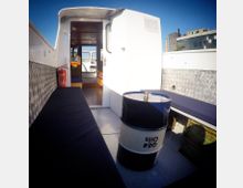 Innenraum eines kleinen Boots mit Blick auf den offenen Durchgang zur Kabine. Die Sitzflächen sind mit dunklen Polstern bedeckt, eine Ölfass-ähnliche Tonne steht in der Mitte, und eine Feuerlöscherhalterung ist an der Wand angebracht. Im Hintergrund sind moderne Gebäude sichtbar.