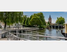 Ein Flussufer mit einer Holzpromenade und einem Geländer im Vordergrund, flankiert von großen Trauerweiden und anderen Bäumen. Im Hintergrund sind ein spitz zulaufender Backsteinturm und ein Gebäude am Wasser zu sehen.