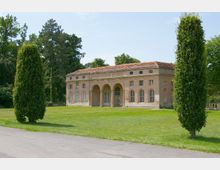 Klassizistisches Gebäude mit einem ziegelroten Walmdach und einer Fassade mit Rundbogenfenstern sowie drei großen Rundbögen am Eingang. Das Gebäude steht in einer gepflegten Parklandschaft mit Rasen, Bäumen und hochgewachsenen Heckenpflanzen im Vordergrund.
