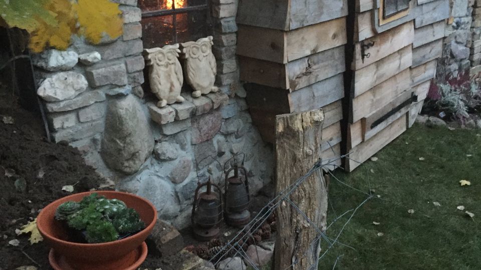 Steinerne Gebäudeecke mit kleinem, vergittertem Fenster und warmem Licht im Inneren, daneben ein holzverkleideter Anbau mit kleiner Tür und Fenster. Im Vordergrund Gartenbereich mit Pflanzschale, Drahtzaun, Holzpfosten und herbstlichen Ahornblättern.
