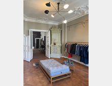 Ein stilvoller Innenraum mit einem verzierten Deckenstuck, einer hellgrünen, antiken Kachelheizung sowie Holzfußboden. Kleidung hängt ordentlich auf einer Kupferstange an der Wand, und in der Mitte steht eine gepolsterte Holzbank. Ein Durchgang führt in einen weiteren Raum.