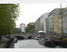 Städtische Szene mit einer Wasserstraße, entlang der Boote festgemacht sind. Rechts stehen mehrstöckige Gebäude mit unterschiedlichen Fassaden, links ein Gehweg mit Bäumen, und im Hintergrund ist eine steinerne Brücke sowie ein modernes Hochhaus sichtbar.