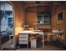 Innenraum eines alten Untersuchungs- oder Labors: ein Schreibtisch mit Lampe und Waage steht vor einer Liege, an der Wand hängen Glasschrank und Leitungen. Rechts befindet sich ein kleines Waschbecken, links ist durch eine Öffnung ein weiterer Raum mit Untersuchungsliege und Wandgerät zu sehen.