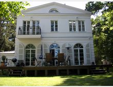 Zweistöckiges weißes Gebäude im klassischen Stil mit großen Fenstern und einem kleinen Balkon. Vor dem Haus befindet sich eine Holzterrasse mit Gartenmöbeln, umgeben von einem gepflegten Garten mit Bäumen.