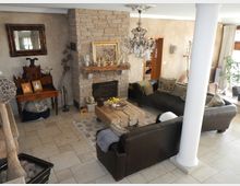 Ein gemütliches Wohnzimmer mit einem Kamin aus Naturstein in der Mitte, flankiert von Ledersofas und einem rustikalen Holztisch. Der Raum ist mit Dekorationen wie einem Kronleuchter, Pflanzen, Bildern und einer antiken Kommode eingerichtet, und helle Fliesen bedecken den Boden.