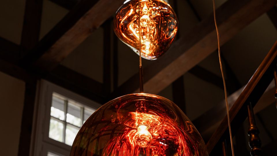 Drei hängende Glas-Pendelleuchten mit warmem, orangefarbenem Licht in einem Innenraum mit sichtbaren Holzbalken und großen, mehrteiligen Fenstern. Im Hintergrund ist ein Treppenaufgang mit dunklem Geländer zu sehen.