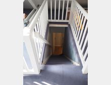 Treppenaufgang mit blauem Teppichboden, umgeben von weißem Geländer. Die Treppe führt in einen unteren Bereich mit Holzboden.