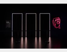 Dunkler Raum mit drei vertikalen, von weißem Licht eingefassten Türöffnungen, die von Vorhängen verdeckt sind. Rechts an der Wand leuchtet eine pinke Neonzeichnung in Form eines stilisierten Gesichts.