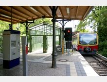Ein Bahnhof mit einem Bahnsteig unter einem hölzernen Dach, einer Fahrkartenmaschine und einem wartenden Zug der Berliner S-Bahn. Im Hintergrund ist ein verglaster Übergang und grünes Laub sichtbar.