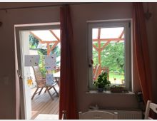 Blick durch eine Terrassentür und ein Fenster mit orangefarbenen Vorhängen in einen Garten mit Holzterrasse und Gartenmöbeln. Auf der Glastür sind Kinderzeichnungen angebracht, und auf der Fensterbank stehen Pflanzen und Schalen.
