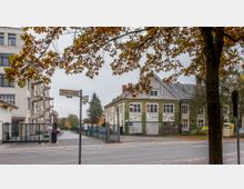 Straßenszene mit herbstlichem Laub im Vordergrund, einem Straßenschild mit den Namen "Goerzallee" und "Albesweg", einem alten Gebäude mit grüner und weißer Fassade rechts sowie modernen Gebäuden links; ein Zaun grenzt einen Zugang mit Tor ab.