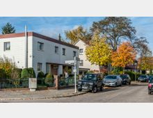 Straßenszene in einer Wohngegend mit einem zweistöckigen weißen Gebäude aus Klinker, einem Vorgarten mit Hecken und einem Zaun. Auf der Straße parken mehrere Autos, und Bäume mit herbstlich gefärbtem Laub säumen den Gehweg.