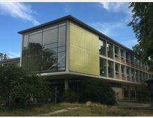 Modernes Schulgebäude mit großen Glasfenstern und einer Fassade aus gelben und roten Kacheln, umgeben von Bäumen und Büschen. Das Gebäude hat zwei Stockwerke und ein Vordach über dem Eingangsbereich.