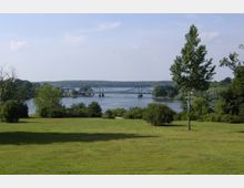 Grüne Parklandschaft mit Blick auf einen breiten Fluss, über den eine filigrane Stahlbrücke mit Bögen führt (die Glienicker Brücke). Im Hintergrund sind Wälder und ein bewölkter Himmel zu sehen.