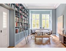 Ein helles Arbeitszimmer mit hohen Decken und großen Fenstern, die Tageslicht hereinlassen. Links befindet sich eine große, eingebaute Bücherwand mit einer Leiter, während in der Mitte ein Tisch mit Stühlen steht; der Boden besteht aus Fischgrätparkett.