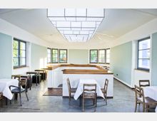 Heller Innenraum eines modernen Restaurants mit weiß gedeckten Tischen und Holzmöbeln. Große Fenster mit schwarzem Rahmen lassen natürliches Licht herein, während eine geometrische Deckenbeleuchtung den Raum dominiert.