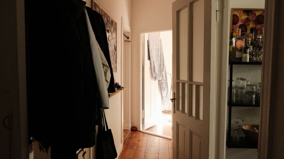 Schmaler Wohnungsflur mit Holzdielen, heller Wand und Garderobe an der linken Seite. Rechts steht eine geöffnete Tür mit Glaseinsätzen neben einem Regal mit Flaschen und Gläsern; am Ende führt eine offene Tür in einen hell erleuchteten Raum.