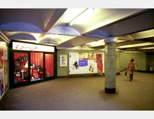 Unterführung mit grün gefliesten Wänden und einem hell beleuchteten Geschäft mit rotem Vorhang und ausgestellten Koffern. An der Wand befinden sich mehrere Plakate, eine Person geht in Richtung eines Ausgangs.