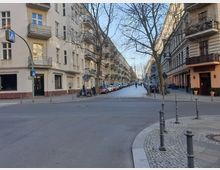 Straßenszene in einer städtischen Umgebung mit Wohnhäusern im Altbaustil links und rechts, teils mit Balkonen. Im Vordergrund eine Straßenkreuzung mit Gehwegen, Pollern und einem Zebrastreifen; parkende Autos und ein Baum reihen sich entlang der gepflasterten Seitenstraße.