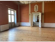 Großer, leerer Saal mit breitem Holzdielenboden, rot gemusterten Wänden und weißer Holzvertäfelung sowie Stuckfries unter der Decke. Links ein hohes Fenster, rechts eine doppelflügelige Tür mit Wappen darüber, die in eine Flucht weiterer Räume führt; ein Kronleuchter hängt von der Decke.