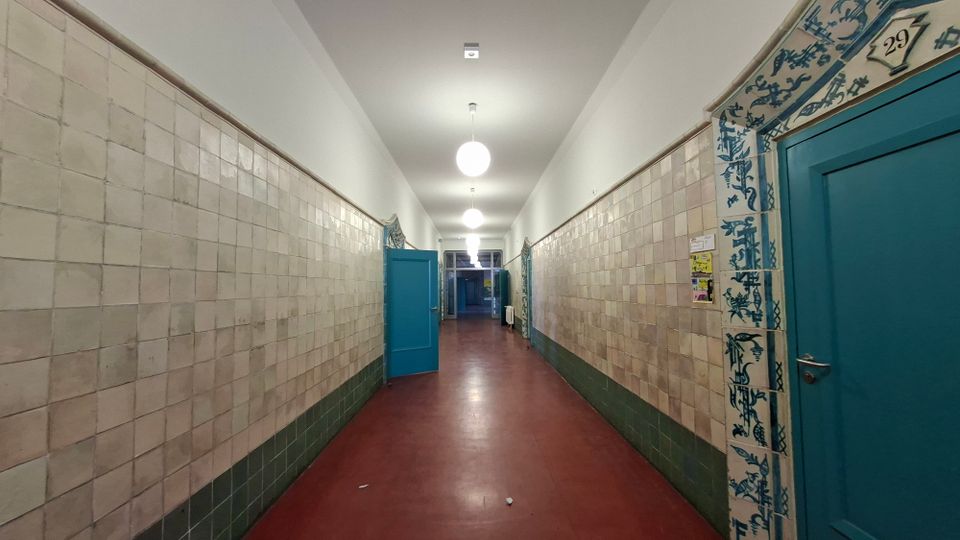 Langer, schmaler Innenflur mit beigefarbenen Wandfliesen und dunkelgrünem Sockelbereich sowie rotem Bodenbelag. Entlang des Gangs befinden sich blaue Türen und runde Deckenleuchten; eine Türzarge ist mit blau-weißem Fliesendekor und einer Nummerntafel verziert.