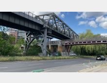 Eine zweistöckige Brücke aus Metall mit Fachwerkträgern, unter der eine Straße verläuft. Auf einem Betonpfeiler steht der Schriftzug „BERLIN“, und die Umgebung ist von Bäumen und Graffiti geprägt.