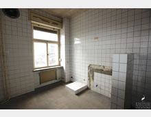 Ein leerer Raum mit weiß gefliesten Wänden und einem grauen Fliesenboden. Ein großes Fenster mit einem Heizkörper darunter sowie ein rechteckiger Ausschnitt in der Wand sind sichtbar; einige Rohre und ein kleiner, gefliester Podest befinden sich ebenfalls im Raum.