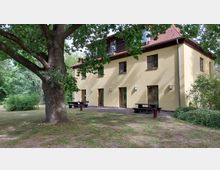 Gelbes zweistöckiges Gebäude mit Satteldach und Holzfenstern in einer grünen, parkähnlichen Umgebung; davor stehen mehrere Holzbänke und ein großer Baum spendet Schatten.