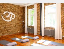 Ein heller Yogaraum mit einer Ziegelwand, weißen Vorhängen an großen Fenstern und einem Holzboden. Matten und Bolster liegen ordentlich auf dem Boden, und an der Wand ist ein großes, weißes Om-Symbol angebracht.