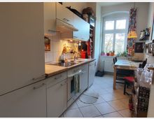 Narrow galley kitchen with gray cabinets, a long countertop with sink and stove, and white tile backsplash. At the far end, a large arched window above a radiator sits beside a small dining table with chairs, patterned curtains, and potted plants on the sill.