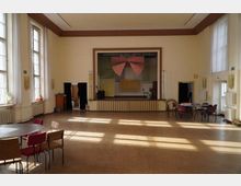 Ein heller Mehrzwecksaal mit hohen Fenstern an beiden Seiten, einer kleinen erhöhten Bühne am Ende des Raumes und mehreren Stühlen und Tischen, die locker im Raum verteilt sind. Die Bühne ist mit einem roten Vorhang dekoriert, die Wände beige gestrichen, und der Raum wird durch Tageslicht erhellt.