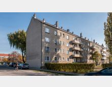 Wohnblock mit mehreren Stockwerken und schlichten, grauen Fassaden. Vorderseite des Gebäudes mit Balkonen und rechteckigen Fenstern; im Vordergrund ein Gehweg, parkende Autos und ein Fahrradfahrer, umrahmt von Bäumen und Hecken.