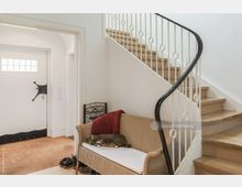 Flur in einem Wohnhaus mit einer hölzernen Treppe, einem weißen Geländer mit runden Elementen und einer Korbbank, auf der eine schlafende Katze liegt. Im Hintergrund befindet sich eine weiße Eingangstür mit einem kleinen Fenster.