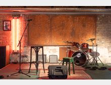 Ein kleiner Bühnenbereich mit einem Schlagzeug, einem Mikrofonständer, einem schwarzen Barhocker und einem grünen Hocker. Die Umgebung zeigt freiliegende Backsteinwände mit Rohren, orangefarbene Lichtelemente und Teppiche auf dem Boden.