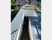 Blick auf eine schmale, außenliegende Treppe, die zwischen weiß verputzten Mauern mit grauen Metallabdeckungen nach unten führt. Oben verläuft ein Geländer entlang einer begrünten Dachfläche; im Hintergrund stehen ein Haus mit Ziegeldach und umgebende Bäume.
