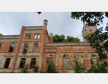 Ruine eines historischen Backsteingebäudes mit teilweise zerstörten Fenstern und Wänden. Im Hintergrund sind Bäume und ein Turm mit Zinnen zu sehen, umgeben von dichter Vegetation.