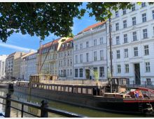 Ein Kanal mit einem alten Holzboot im Vordergrund, umgeben von historischen Gebäudezeilen mit weißen Fassaden und roten Dächern. Im Hintergrund ist ein sonniger, klarer Himmel zu sehen, und Baumzweige rahmen das Bild oben ein.