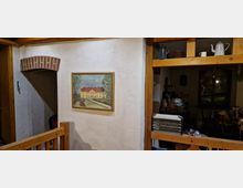Ein Innenraum mit rustikalem Holzrahmen und Putzwänden, auf denen ein gerahmtes Gemälde eines Hauses hängt. Rechts befinden sich offene Holzregale mit Krügen und Töpfen sowie ein Teil eines unaufgeräumten Zimmers im Hintergrund.