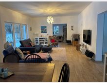 Helles, modernes Wohnzimmer mit großen Fenstern zur verschneiten Außenansicht, grauem Ecksofa mit Ottoman, Teppich und Couchtisch sowie einer auffälligen Kugel-Deckenleuchte. Rechts hängen ein wandmontierter Fernseher und ein kleines Lowboard; im Hintergrund führen Durchgänge in weitere Räume mit Sesseln und Regalen.