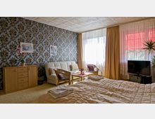 Schlafzimmer in einem Retro-Stil mit geblümter Tapete, einer beigefarbenen Couch, einem Fernseher auf einem schwarzen Tisch, einem Doppelbett mit heller Bettwäsche und einem Couchtisch mit Blumen. Große Fenster mit weißen Vorhängen und gelben Gardinen durchfluten den Raum mit Tageslicht.